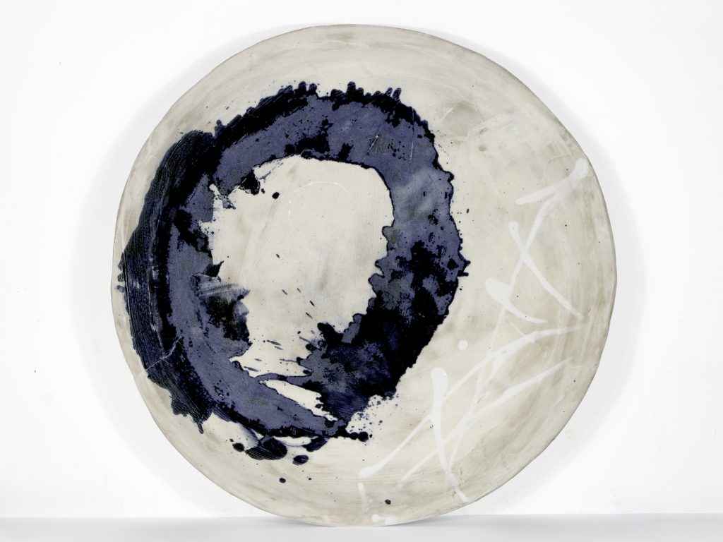 Photograph of large hand-made ceramic plate with grey background glaze, hand-decorated with white glaze splashes and bold dark blue swirl. Artist: Susan Feldman.